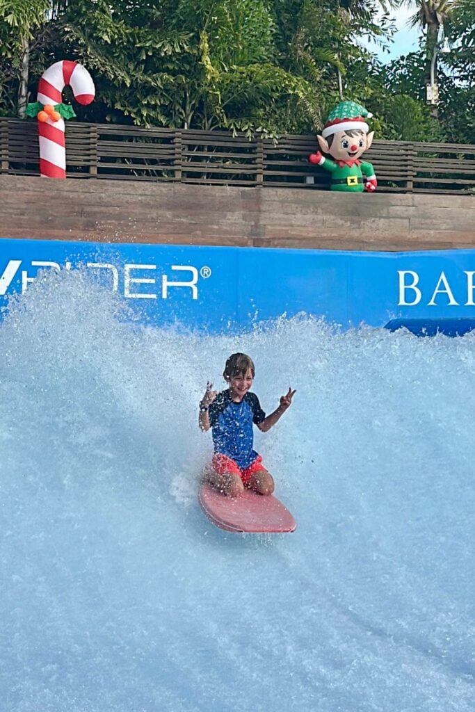 A young boy bodyboarding on the FlowRider surf simulator at Baha Bay water park, Grand Hyatt Baha Mar, flashing a peace sign with Christmas decorations visible in the background.