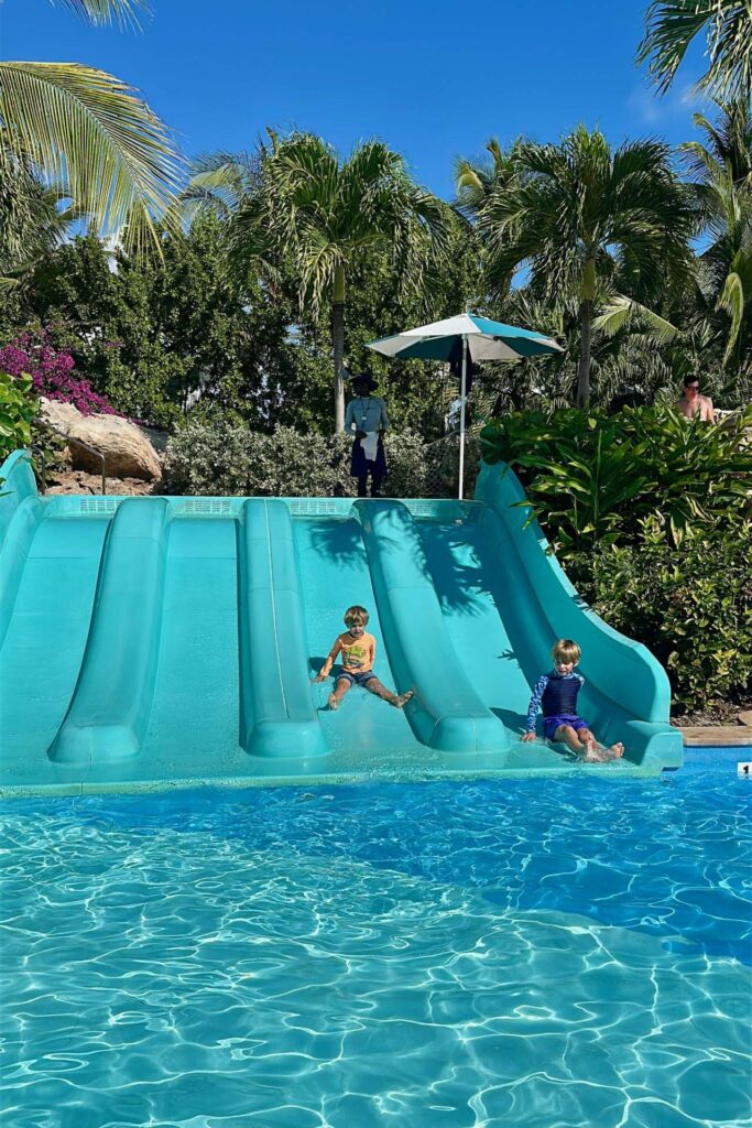 Two young boys racing down side-by-side teal water slides into a bright blue pool at Grand Hyatt Baha Mar, Nassau, Bahamas.