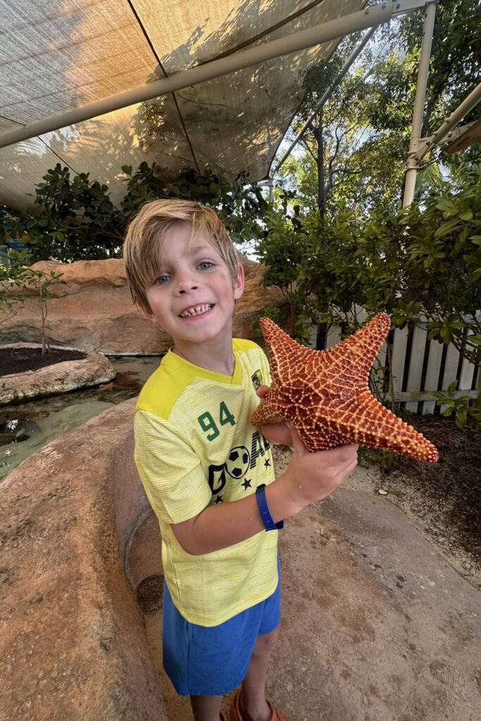 Young boy smiling and holding a large starfish at the Baha Mar Sanctuary touch tank experience in Nassau, Bahamas.