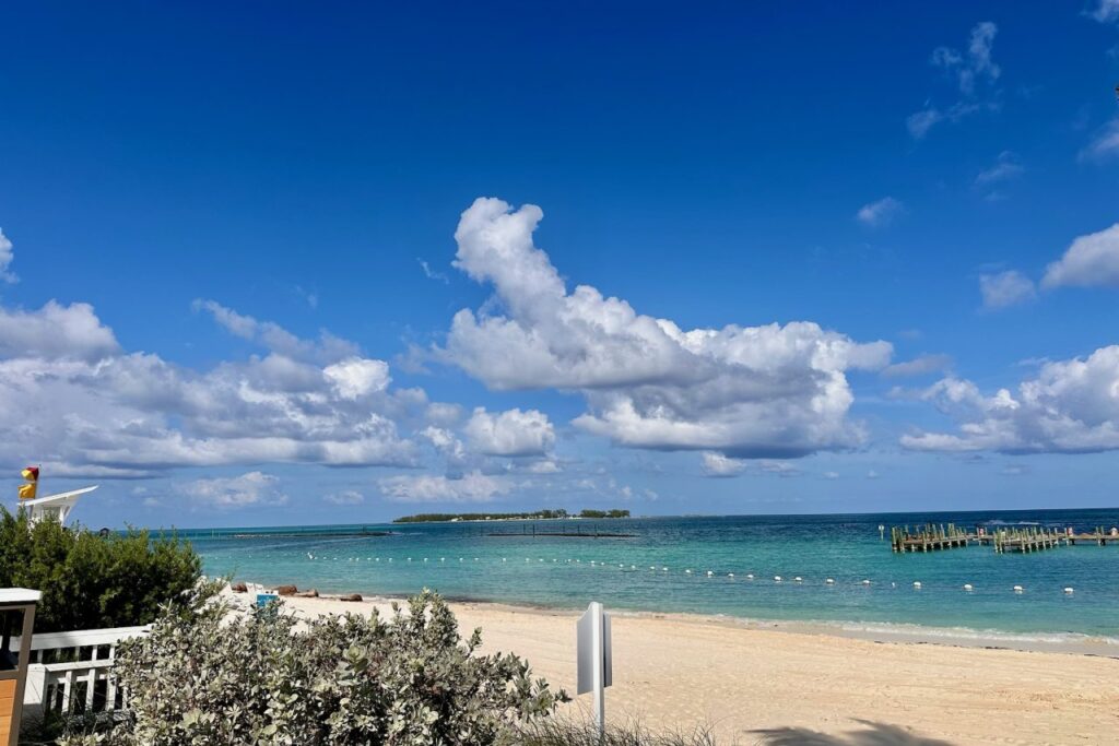 Wide view of Cable Beach at Baha Mar on a clear blue-sky day, showing calm turquoise water, white sand, a wooden pier, and a small island visible in the distance.