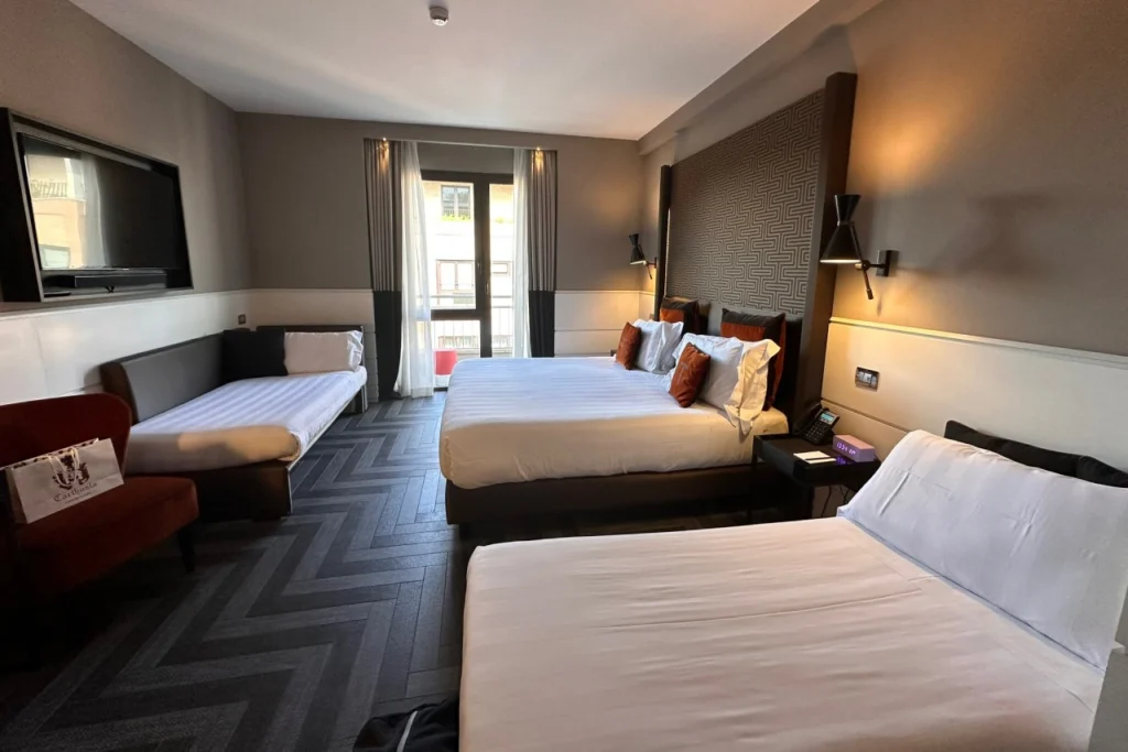 Spacious hotel room at The Tribune Hotel in Rome with three beds, herringbone-pattern dark flooring, warm lighting, and a balcony door