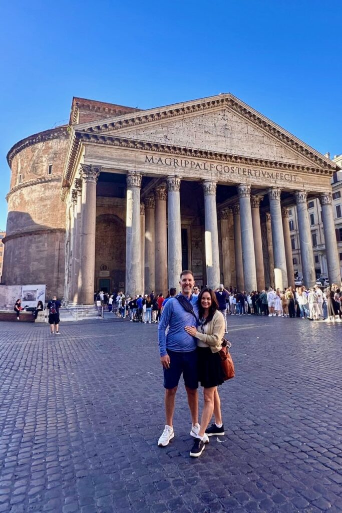 Couple posing in front of the Pantheon in Rome on a clear blue-sky day, with Corinthian columns and the Latin inscription visible behind them