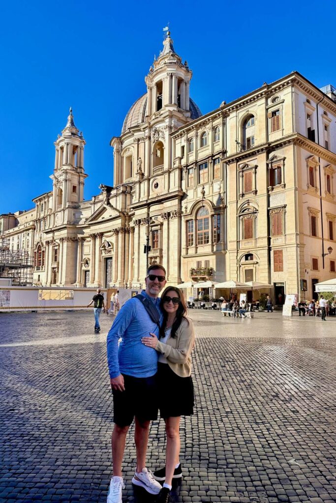 Couple posing in Piazza Navona in Rome on a sunny morning, with the ornate Baroque facade of Sant'Agnese in Agone church behind them
