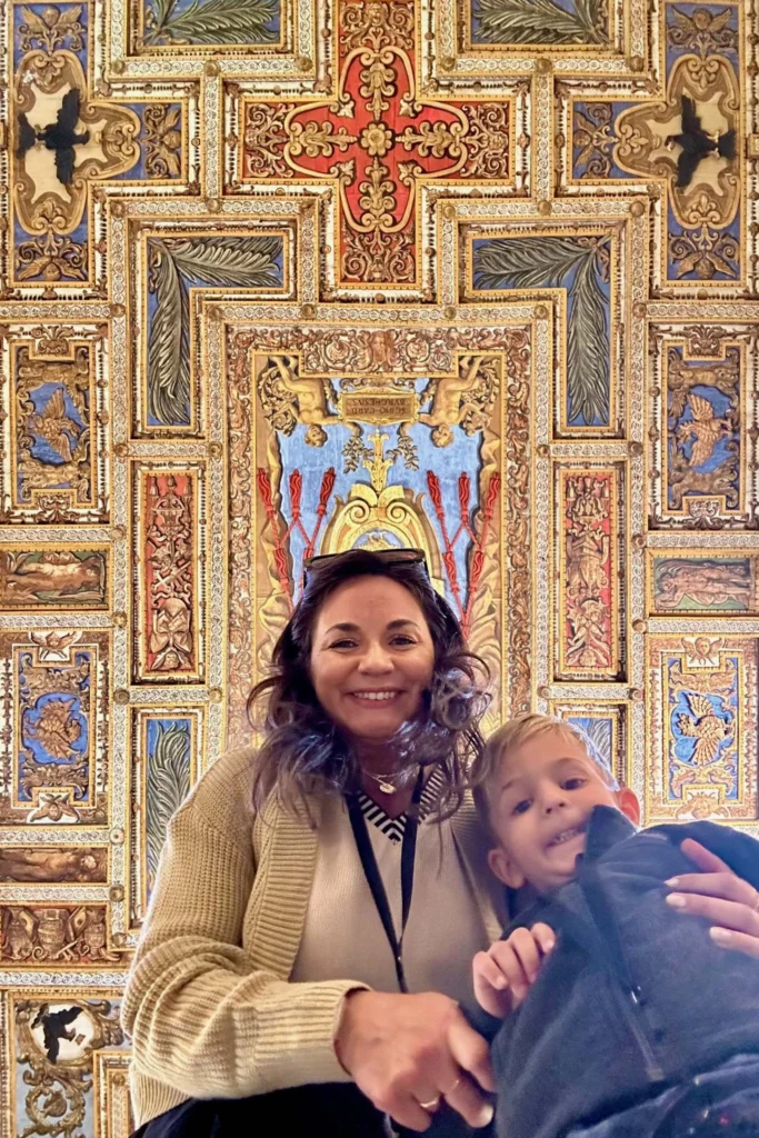 Mother smiling and holding her young son inside the Vatican Museums, with an elaborately decorated gold and painted ceiling visible behind them