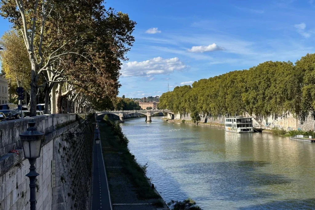 View along the Tiber River in Rome on a sunny autumn day, with tree-lined stone embankments, a bridge in the distance, and a moored river boat