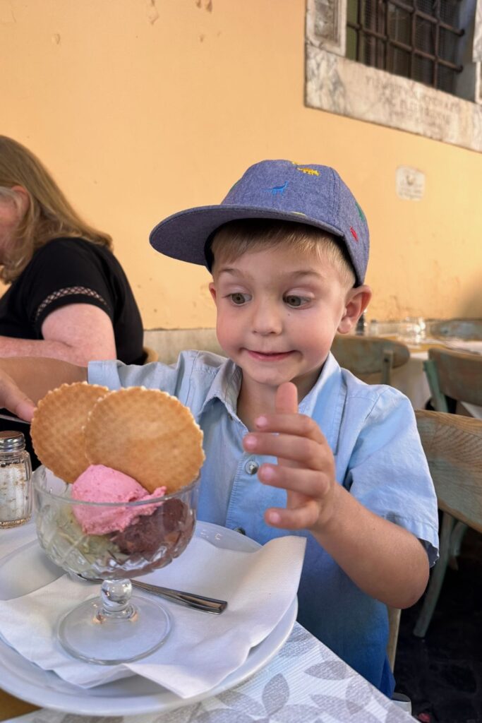 Young boy in a blue cap smiling at a gelato sundae with waffle cookies at a restaurant in Rome
