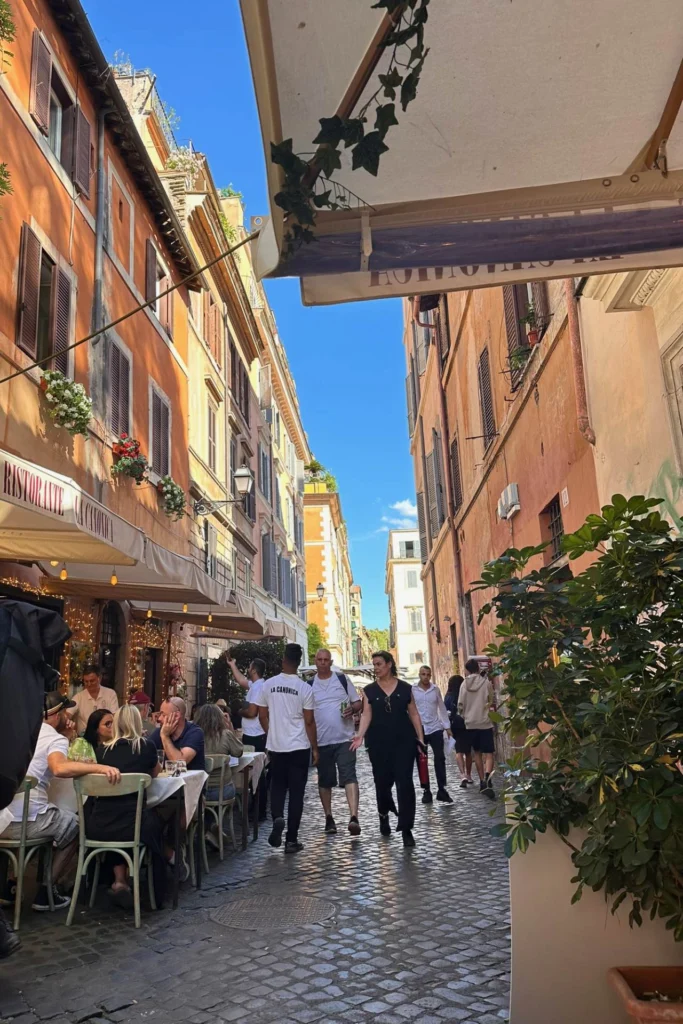 Cobblestone alley in Trastevere, Rome, lined with orange and terracotta buildings, outdoor restaurant seating, and ivy hanging from an awning above