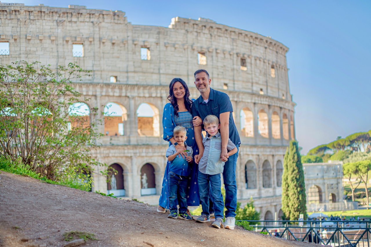 Family of four posing in front of the Colosseum at golden hour, with the mother in a blue floral dress and two young boys standing in front of their parents