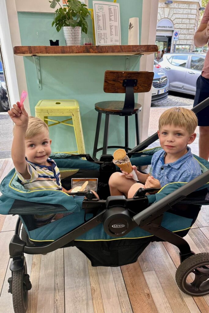 Two young boys sitting in a Graco double stroller eating gelato cones at a gelateria in Rome, one waving his spoon at the camera