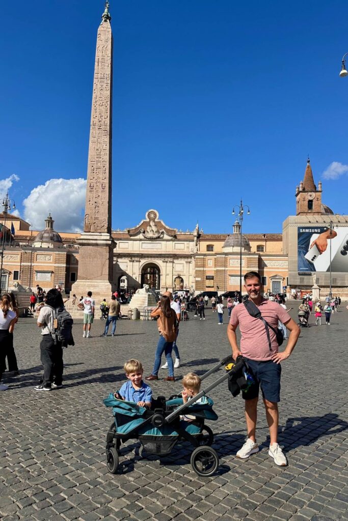 Dad standing with two young boys in a double stroller in Piazza del Popolo, Rome, with the ancient Egyptian obelisk and church facades visible behind them on a sunny day