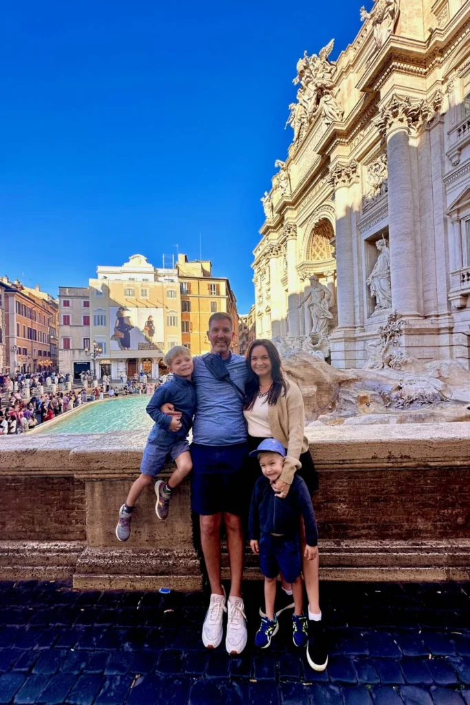 Family of four posing at the Trevi Fountain in Rome, with the turquoise water and ornate Baroque facade and sculptures visible behind them on a bright sunny day