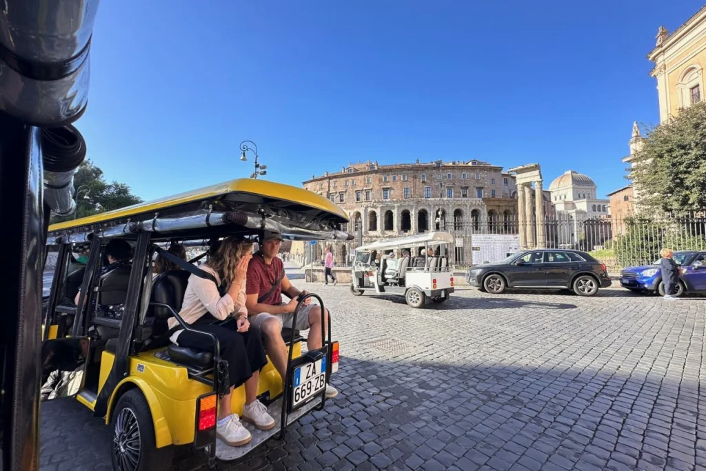 Passengers riding in a yellow golf cart on a cobblestone street during a city tour in Rome, with the Theatre of Marcellus visible in the background on a clear day