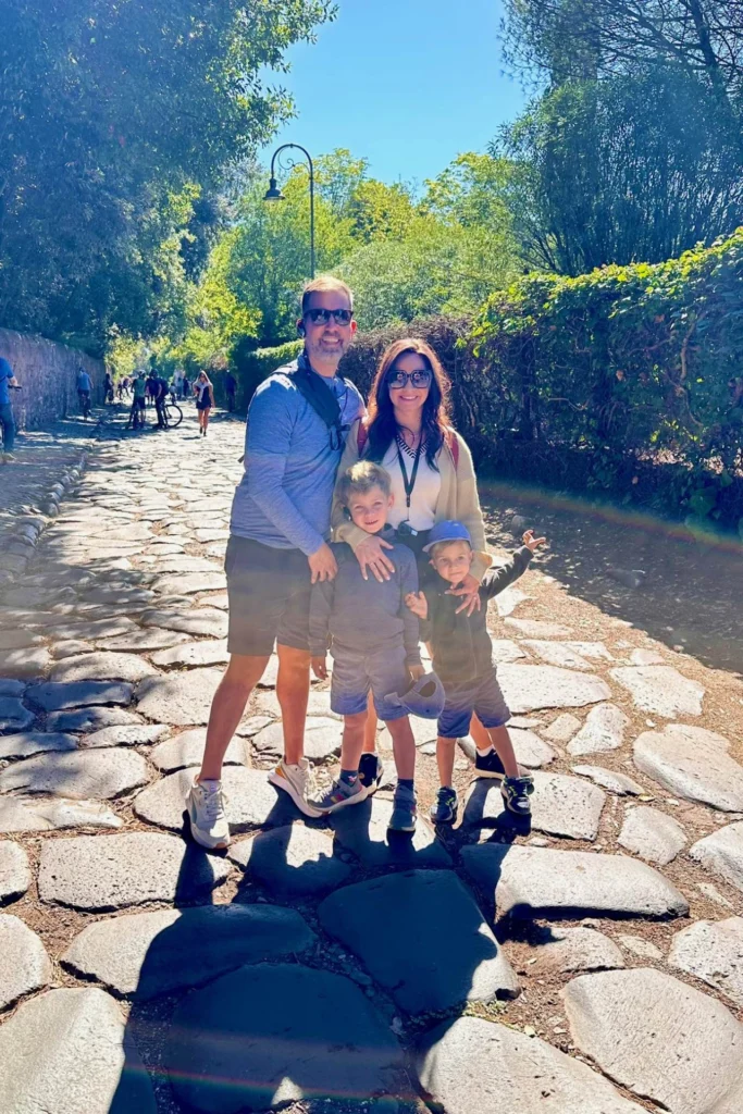 Family of four posing on the ancient cobblestones of the Appian Way in Rome, surrounded by lush greenery and trees on a sunny day, with cyclists visible in the background. things to do in Rome
