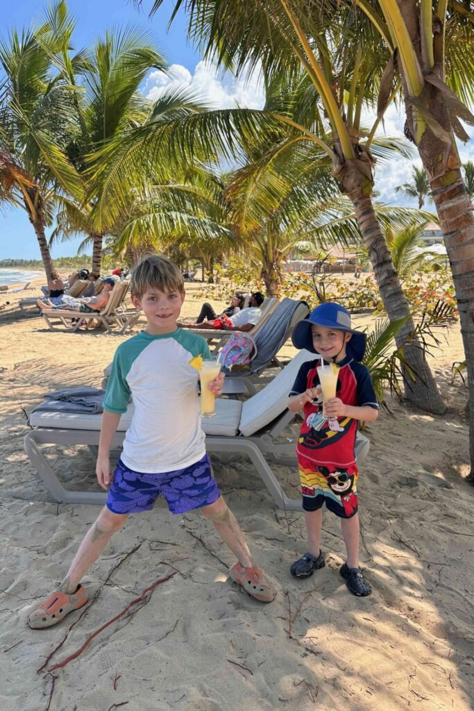 Two boys holding tropical juice drinks on the beach at Dreams Playa Esmeralda under palm trees