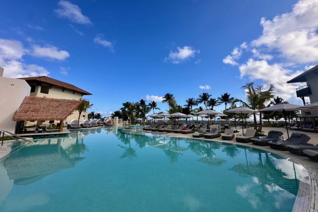 Large resort pool at Dreams Playa Esmeralda on a sunny day with lounge chairs, umbrellas, palm trees, and ocean view beyond