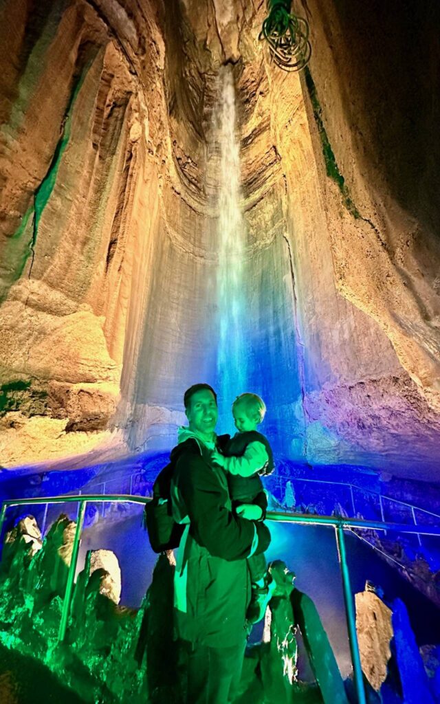 Dad holding a toddler in front of the colorfully lit Ruby Falls waterfall on Lookout Mountain