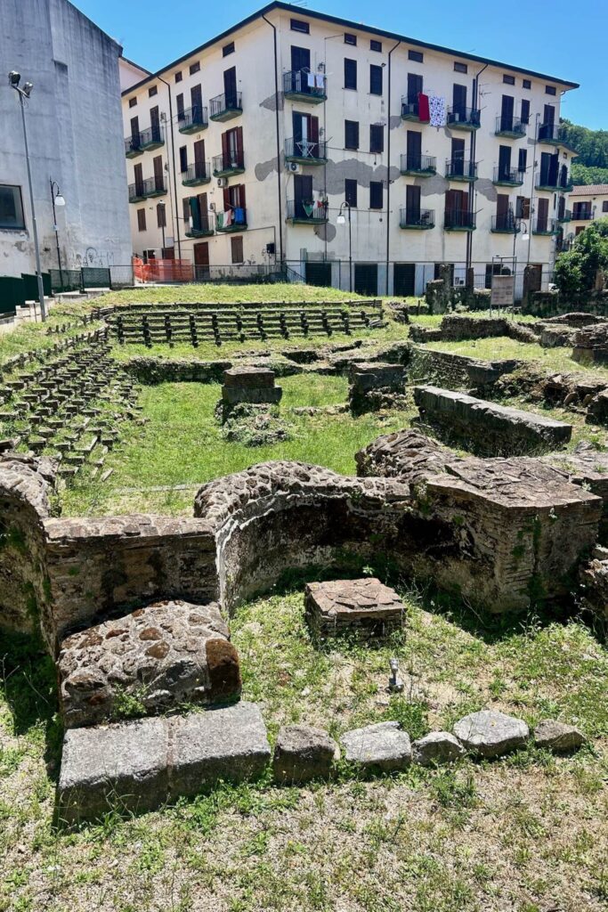 Ancient Roman ruins with stone foundations and tiered seating in Atripalda, set against a backdrop of modern apartment buildings