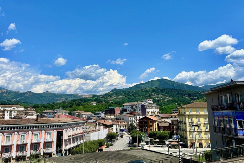 View across an Avellino town center toward forested mountains, with pastel buildings, a piazza, and a statue visible below
