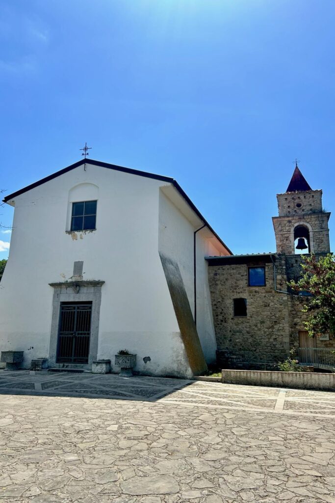 Whitewashed village church with a stone bell tower and bell, fronted by a patterned stone-paved piazza in Santa Paolina