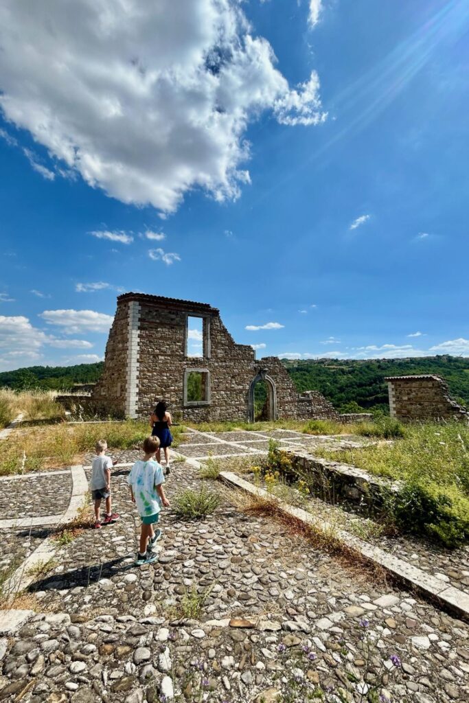 A mother and her two young sons walk through the grassy stone ruins of the old earthquake-destroyed town of Aquilonia, with rolling green hills beyond