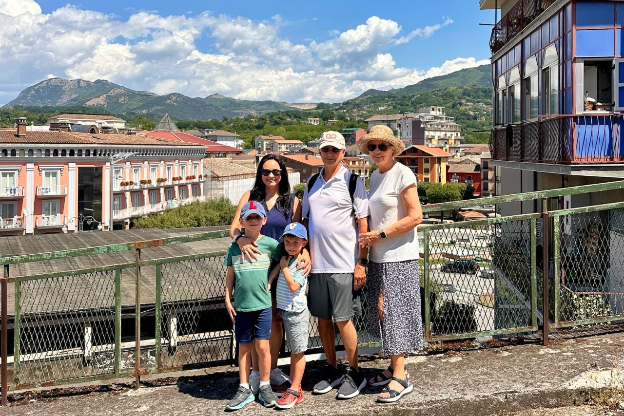 The family poses together on a town overlook, with pastel buildings and green mountains behind them in the Avellino countryside