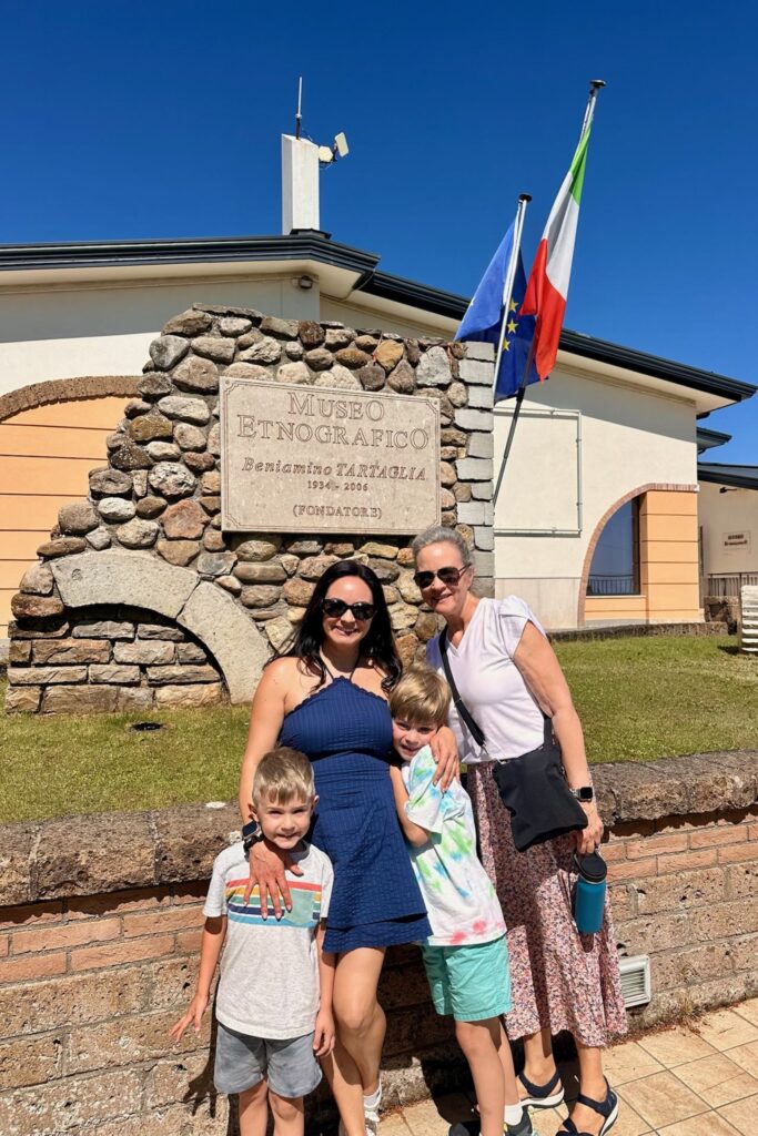 The family poses in front of the Museo Etnografico Beniamino Tartaglia sign in Aquilonia, with Italian and EU flags flying beside the stone entrance