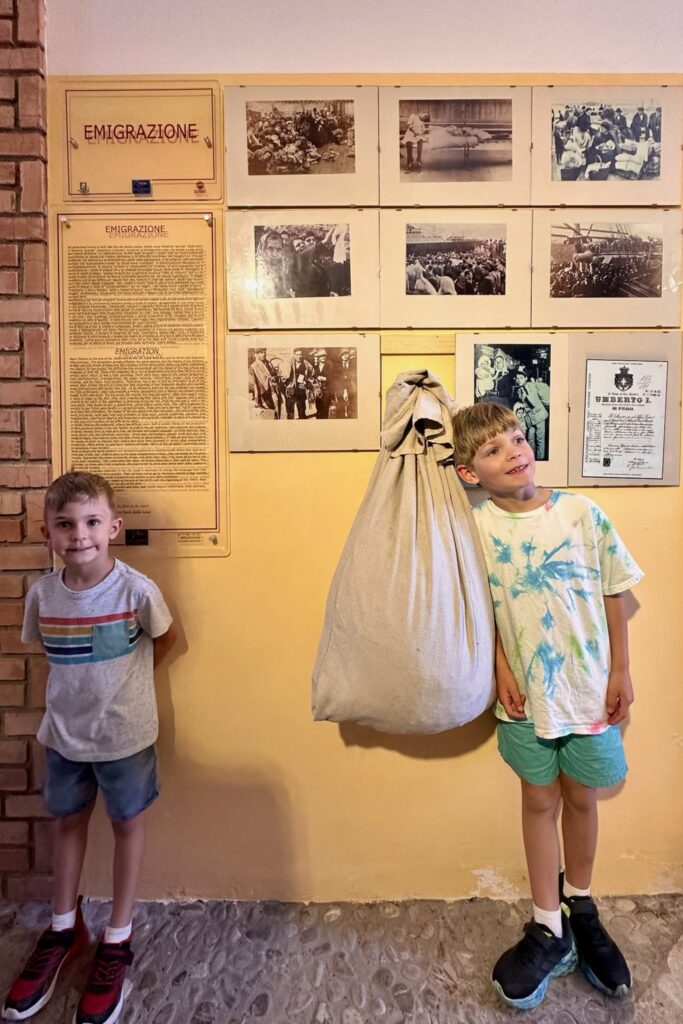 Two brothers stand beside the "Emigrazione" exhibit at the Aquilonia ethnographic museum, next to historical emigration photos and a vintage cloth travel sack