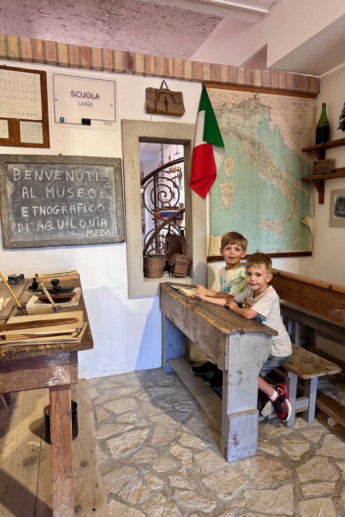 The two boys sit at an antique wooden school desk in the recreated classroom of the Aquilonia ethnographic museum, with a vintage map of Italy and Italian flag on the wall