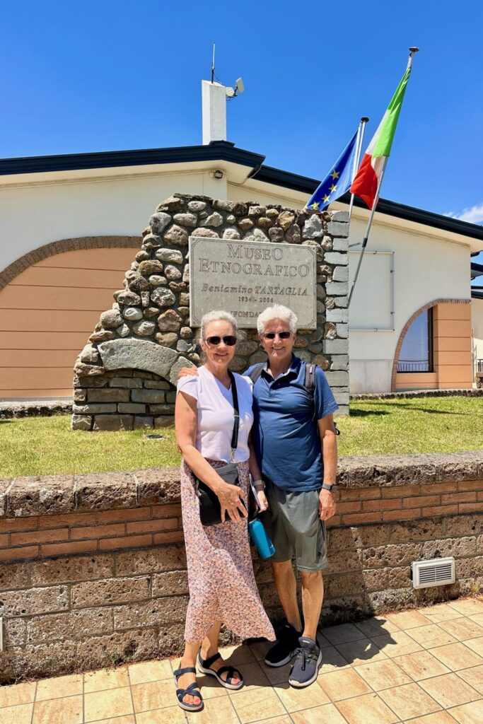 The grandparents stand in front of the stone Museo Etnografico Beniamino Tartaglia entrance sign in Aquilonia, with Italian and EU flags overhead