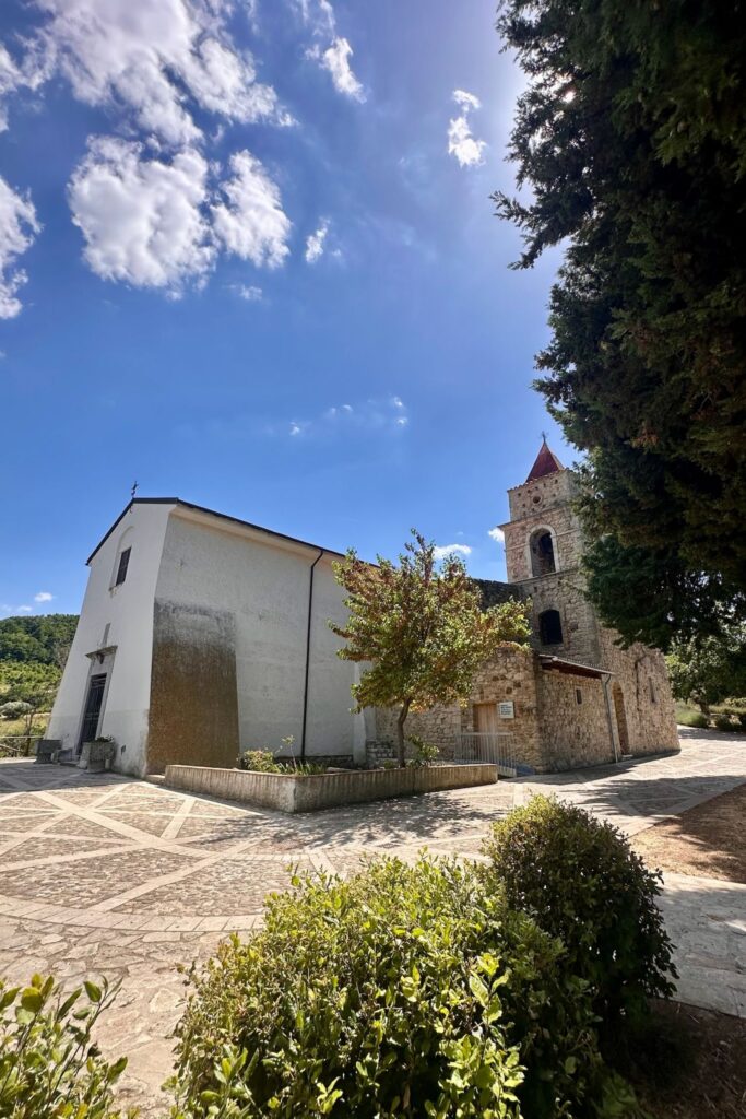 Whitewashed village church with a stone bell tower topped by a red spire, set on a patterned stone piazza in Santa Paolina