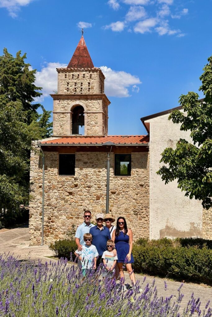 The full family of six poses among purple lavender in front of a stone church and bell tower, with one of the dads joining the grandparents, mom, and two boys