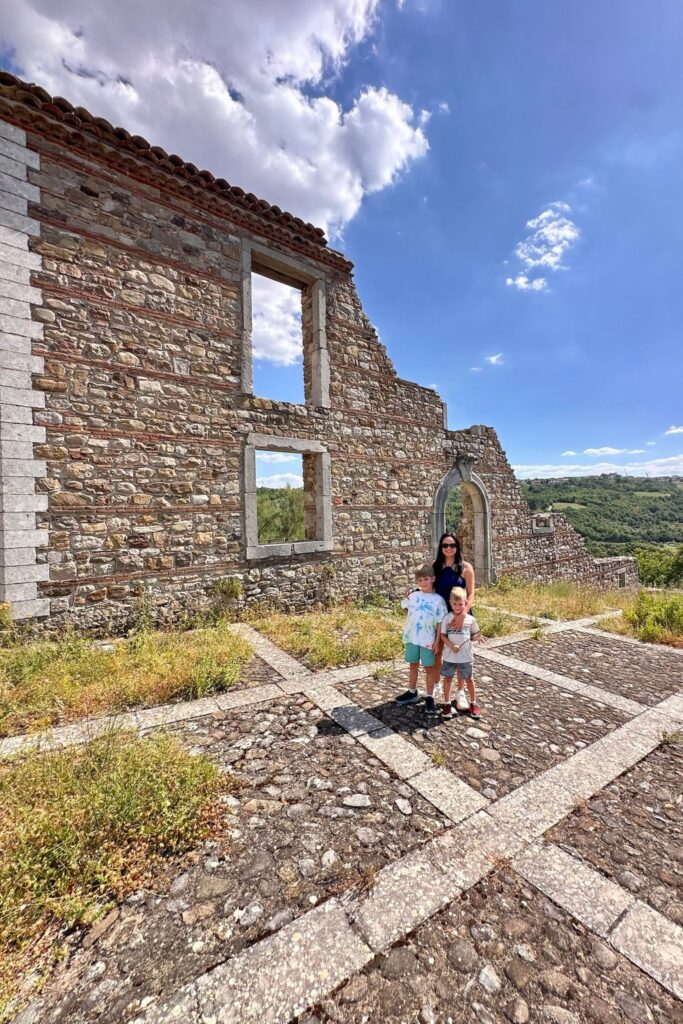 Mom and her two sons stand in the grassy stone ruins of old Aquilonia, with empty window openings, a pointed archway, and green hills behind them