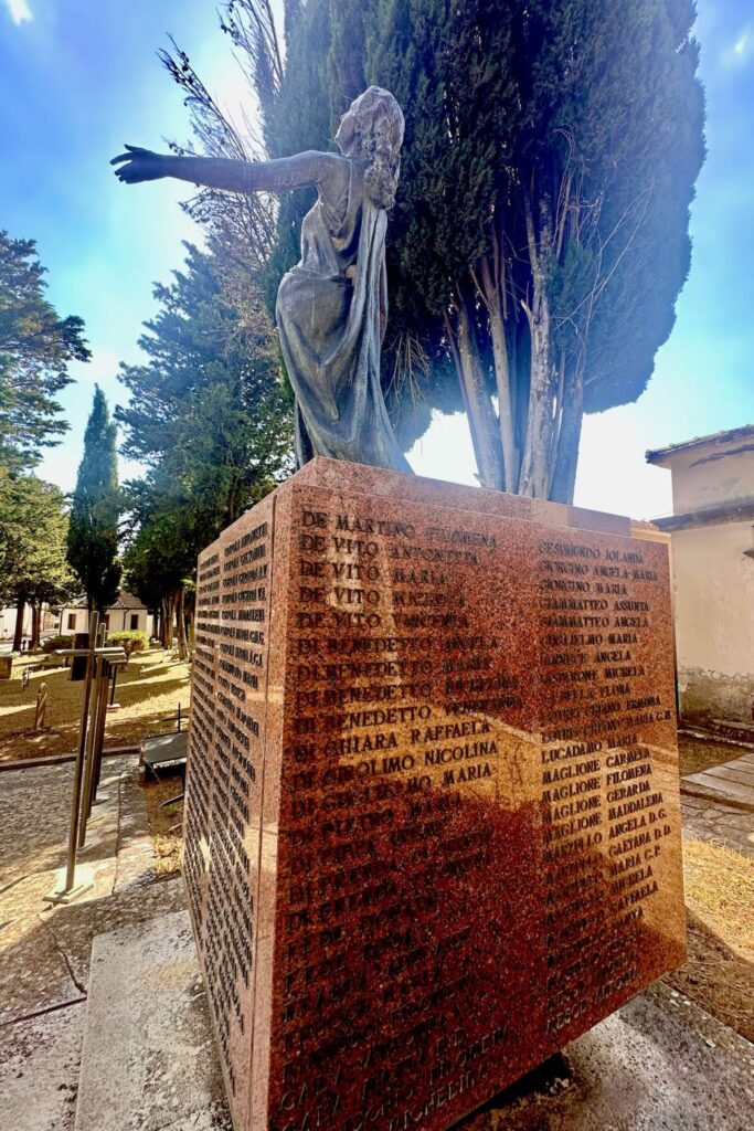 Bronze memorial statue of a robed figure atop a granite plinth engraved with the names of earthquake victims, in the Aquilonia cemetery surrounded by cypress trees