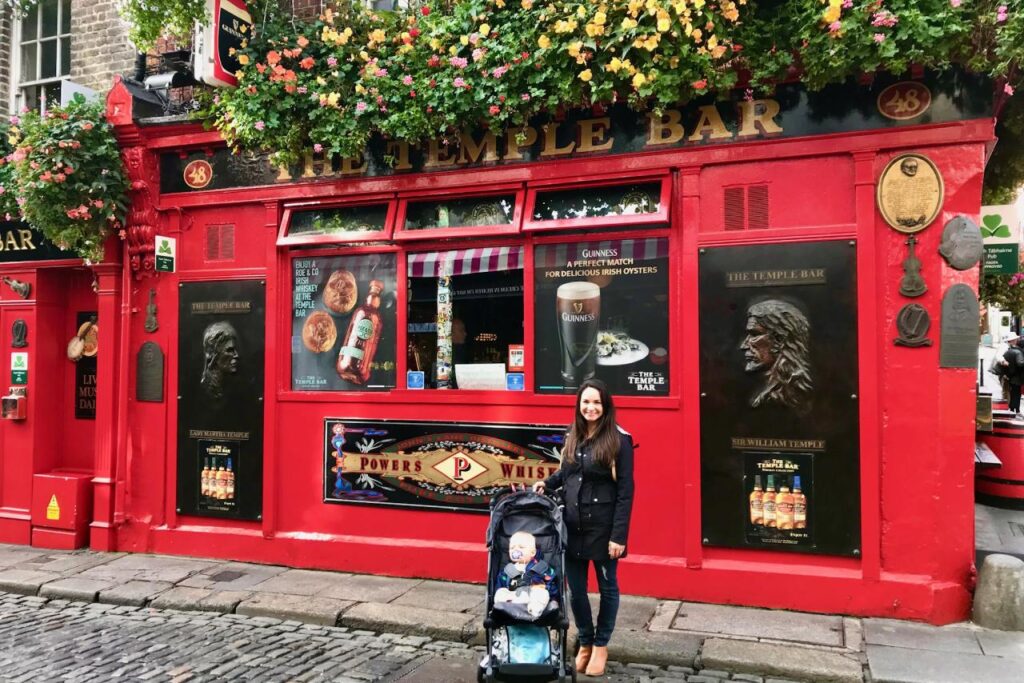 Mom pushing a stroller on the cobblestones in front of the red facade of The Temple Bar pub in Dublin, Ireland.