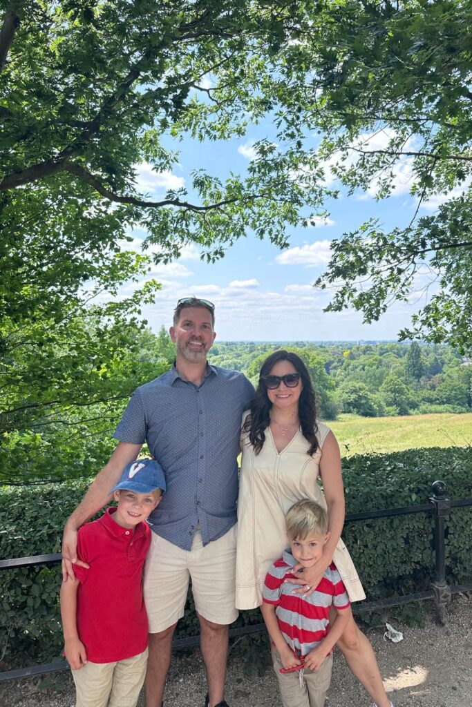 Family of four beneath an oak tree at a Richmond viewpoint with the Thames valley spread out behind them