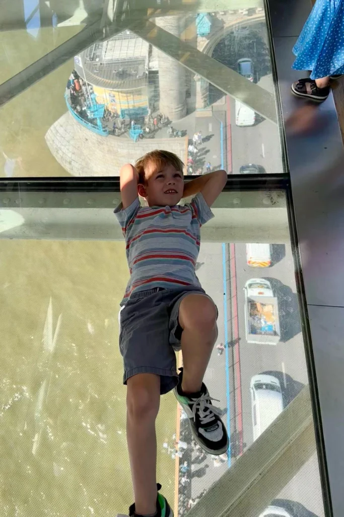 Boy lying on the glass floor of the Tower Bridge walkway with traffic and pedestrians visible below.