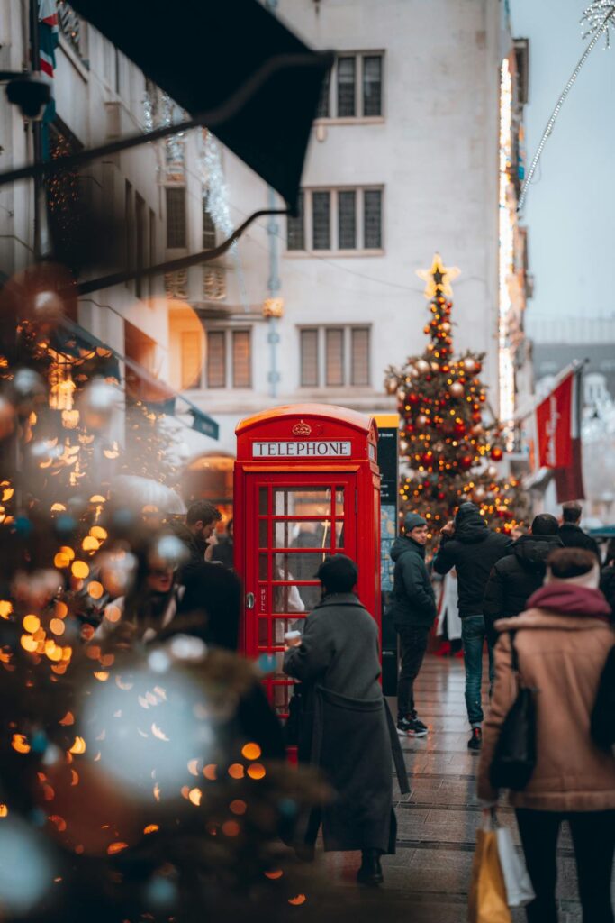 Red telephone box on a London street at Christmas with a decorated tree, shop lights and shoppers walking past.