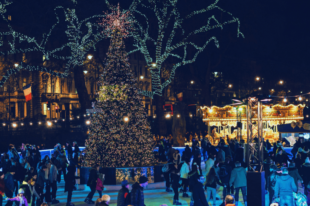 Families ice skating around a lit Christmas tree at an outdoor rink in London, with a carousel glowing behind them.