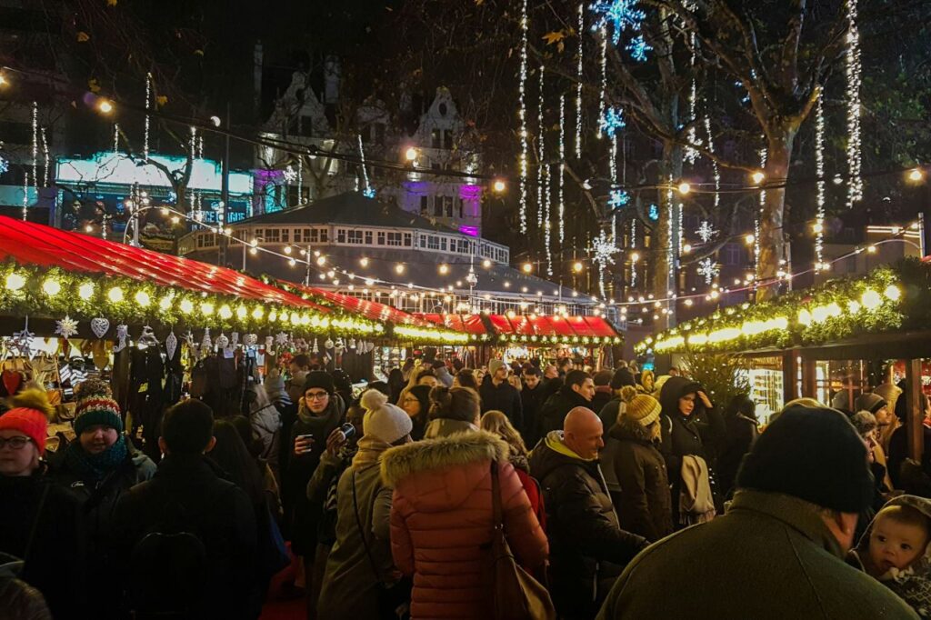 Crowds walking through the Christmas market in Leicester Square at night, with lit stalls and hanging decorations.
