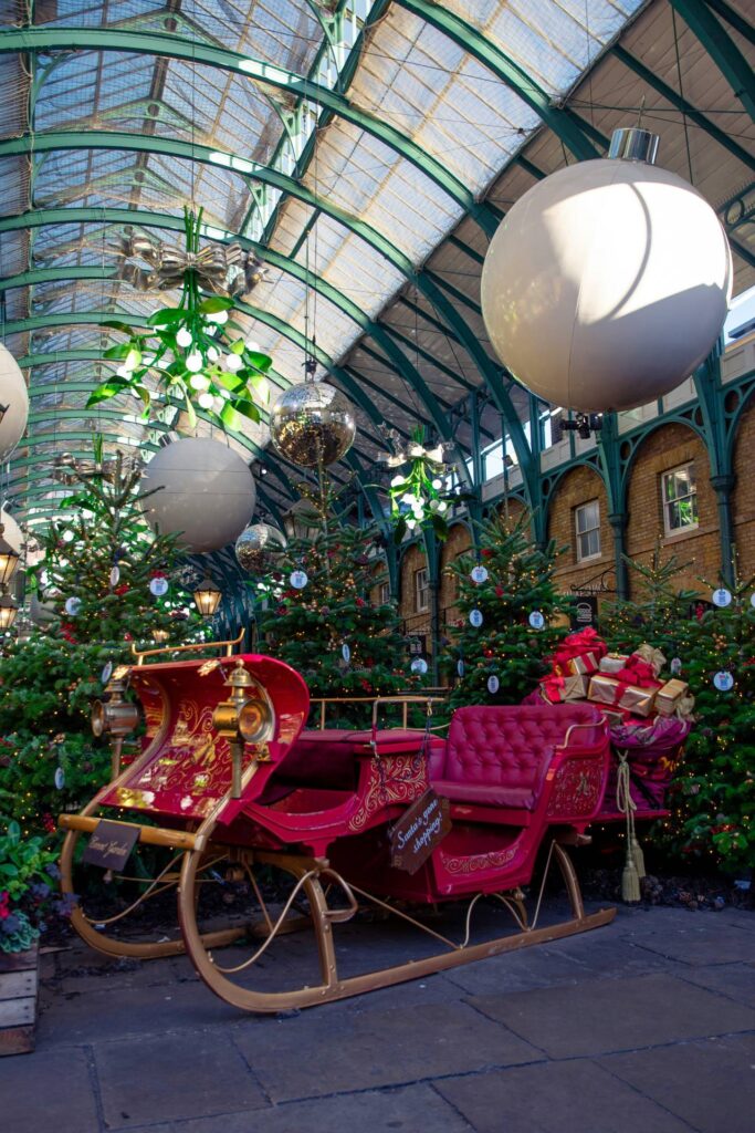 Red and gold sleigh surrounded by Christmas trees and giant baubles inside the Covent Garden market building.