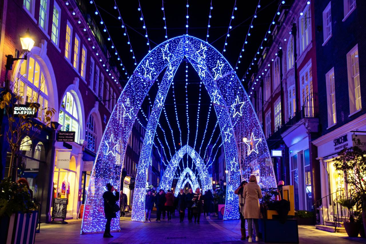 Blue and white star light arches stretching over a pedestrian street in London at Christmas, with people walking beneath.