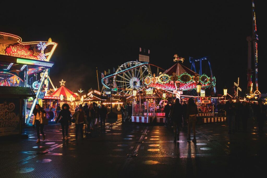 Fairground rides and lit stalls at Hyde Park Winter Wonderland on a wet evening, with a carousel and rollercoaster.
