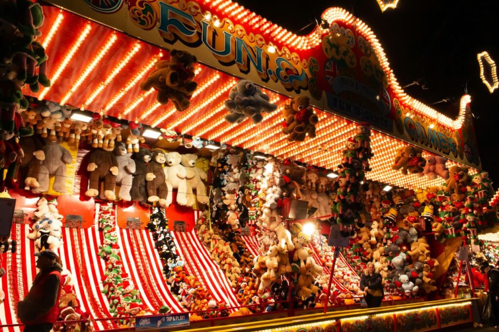 Fairground games stall at Winter Wonderland packed with stuffed toy prizes under bright bulb lighting.