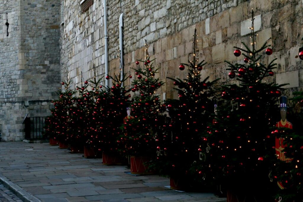 Row of decorated Christmas trees along the stone walls of the Tower of London, with painted Yeoman Warder figures between them.