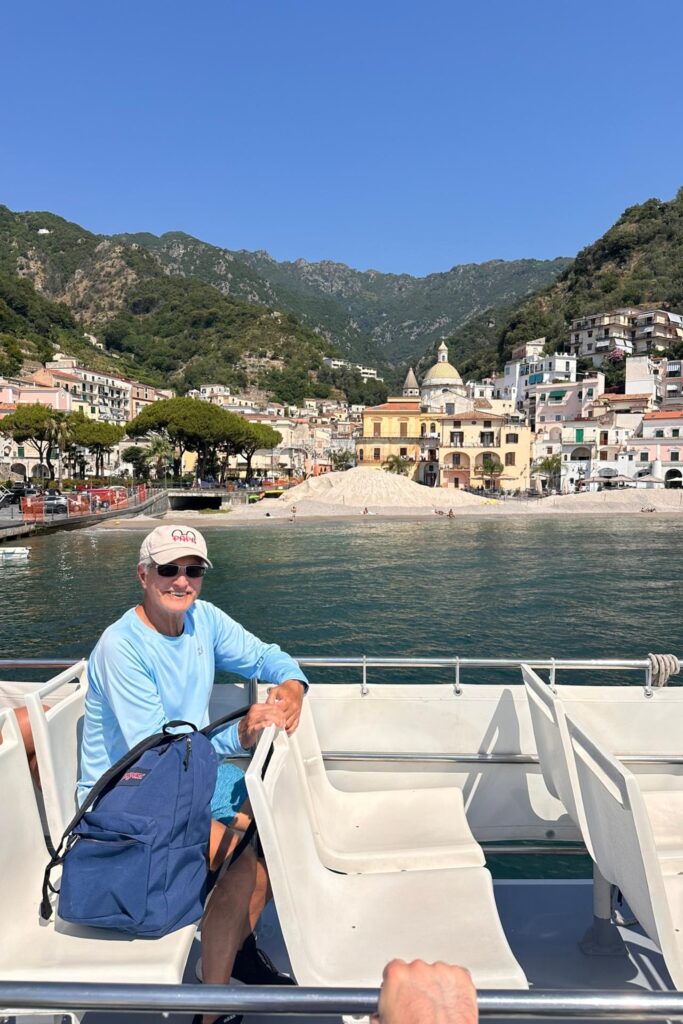 Man smiling on a ferry as it pulls away from Cetara, with pastel houses and a tiled church dome behind him