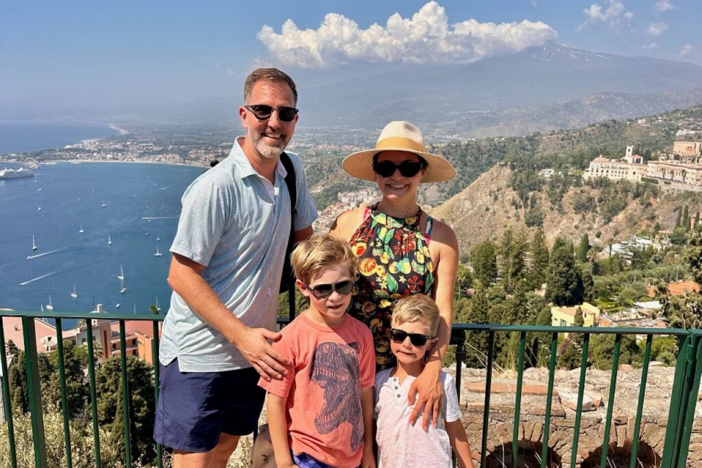 Family of four smiling at a viewpoint in Taormina, Sicily, with sailboats in the bay and Mount Etna in the clouds