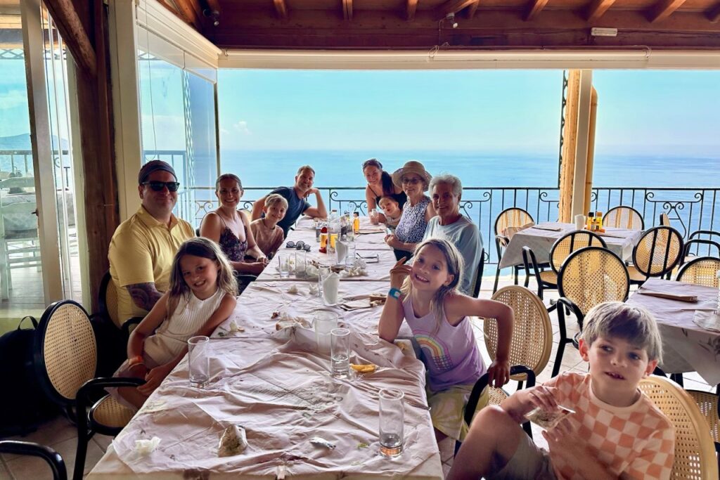 Multigenerational family gathered around a long lunch table on a restaurant terrace with views of the open sea
