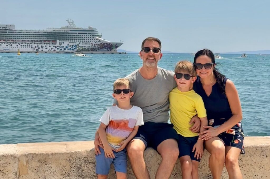 Family of four sitting on a seawall with the Norwegian Gem cruise ship docked in the harbor behind them