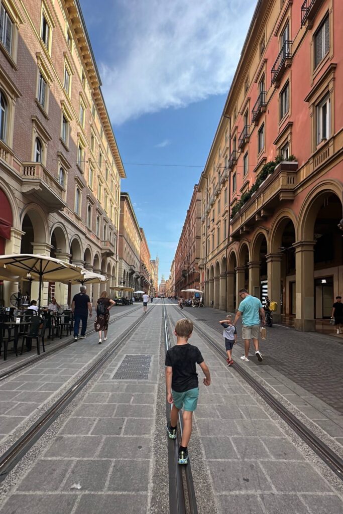 Boys walking along tram tracks on a wide street lined with arched porticoes and terracotta buildings in Bologna