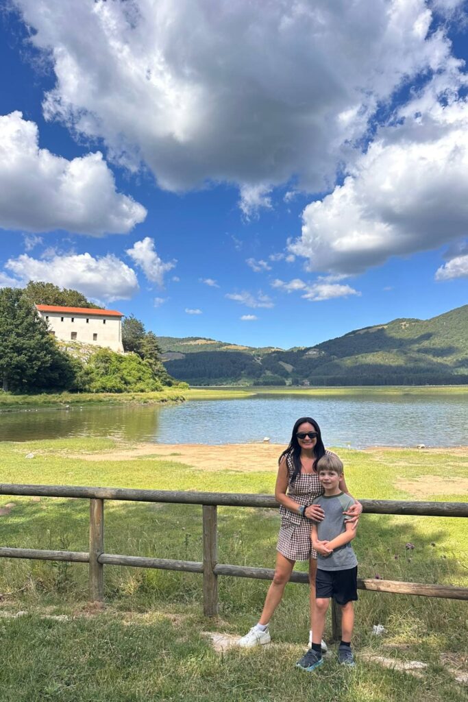 Mom and her son standing by a wooden fence at a mountain lake with forested hills and big white clouds